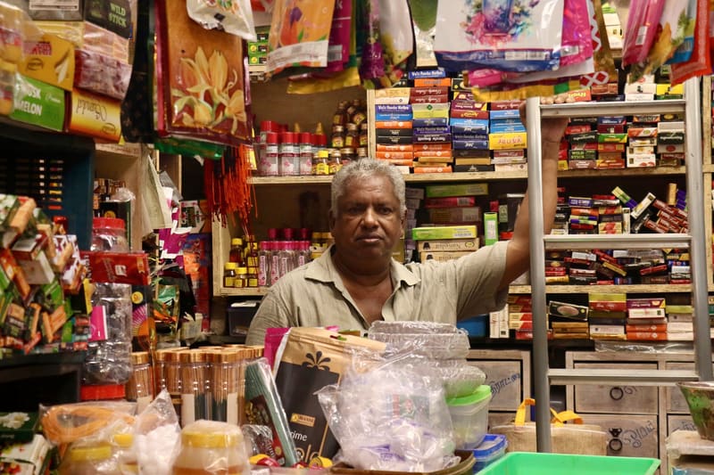 A shopkeeper at the counter of a neighbourhood store, shelves stacked behind him